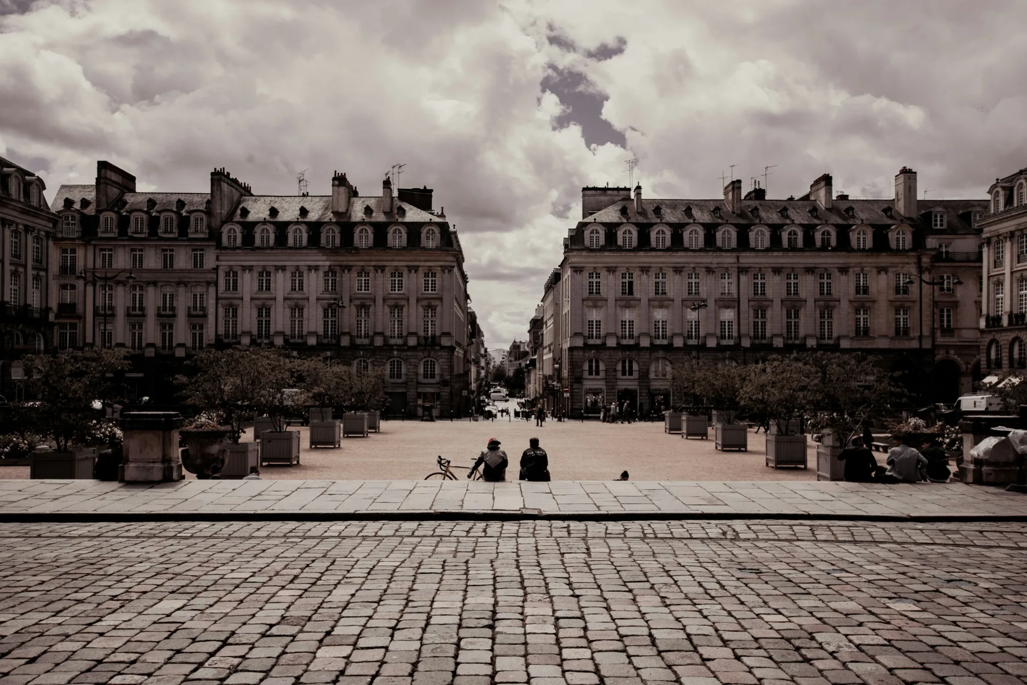 Deux amis assis sur les pavés d'une place du centre de Rennes, face aux façades classiques, ciel nuageux